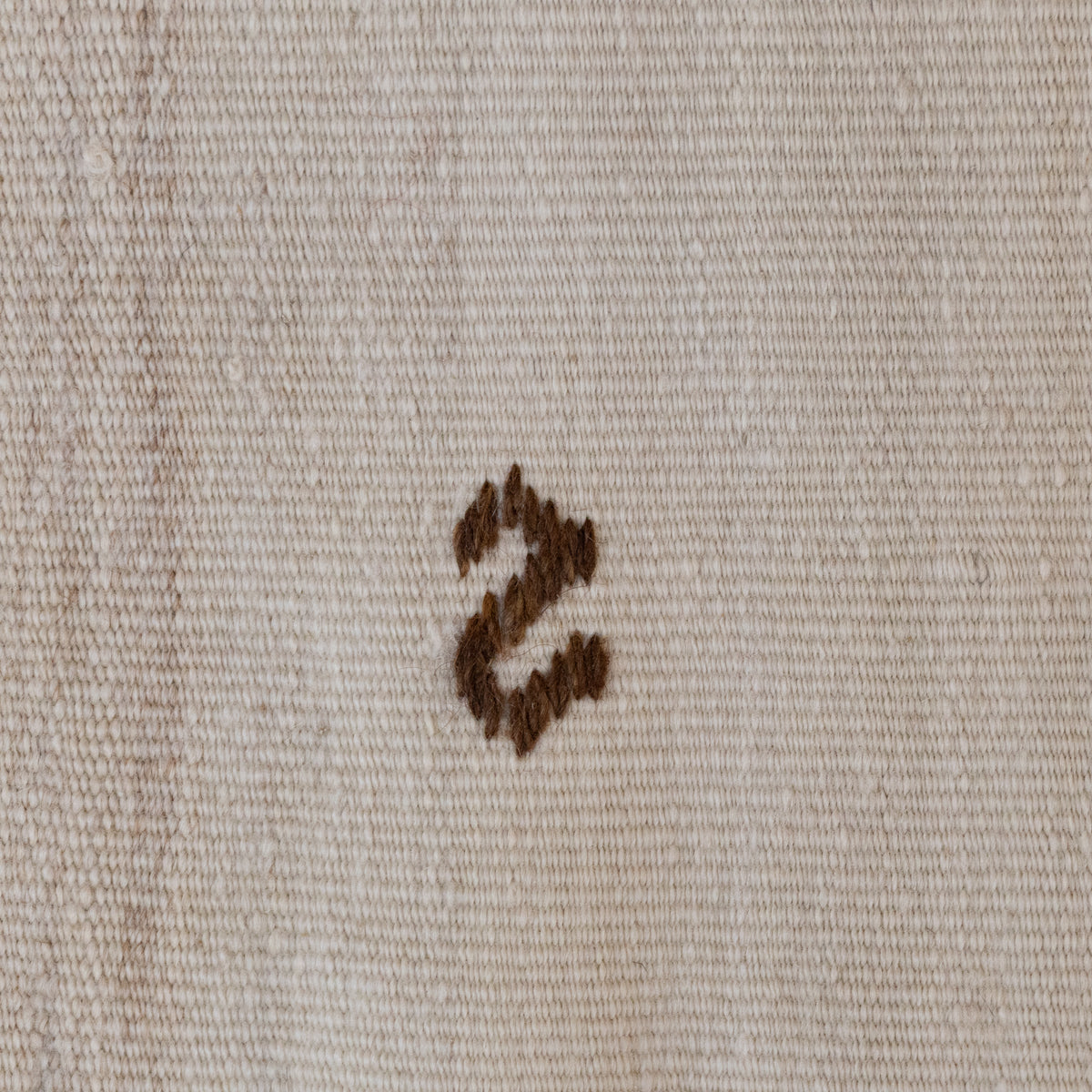 A close-up of a beige woven fabric with a small dark brown embroidered letter 's' in the center.