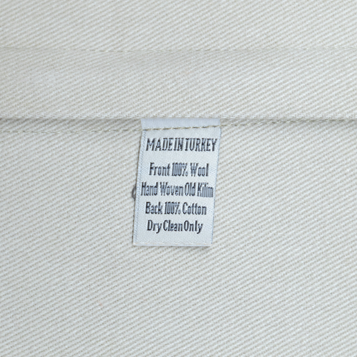 A close-up of a white woven fabric label sewn onto a light gray or off-white textile, showing text that reads 'MADE IN TURKEY', 'Front 100% Wool', 'Hand Woven Old Kilim', 'Back 100% Cotton', and 'DryCleanOnly'.