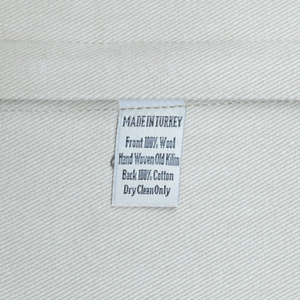 A close-up of a white woven fabric label sewn onto a light gray or off-white textile, showing text that reads 'MADE IN TURKEY', 'Front 100% Wool', 'Hand Woven Old Kilim', 'Back 100% Cotton', and 'DryCleanOnly'.