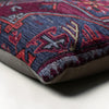 Close-up of a patterned throw pillow corner showing a detailed woven design in deep blue, burgundy red, and beige, with a neutral gray fabric backing visible at the edge.