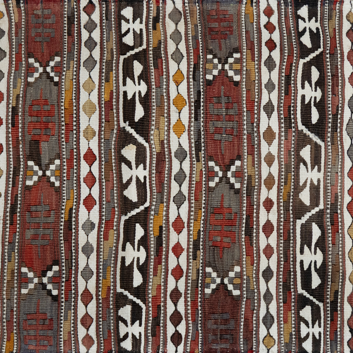 A close-up of a woven textile with vertical tribal-style stripes in red, brown, black, white, gray, and muted yellow, showing detailed fabric texture and pattern.