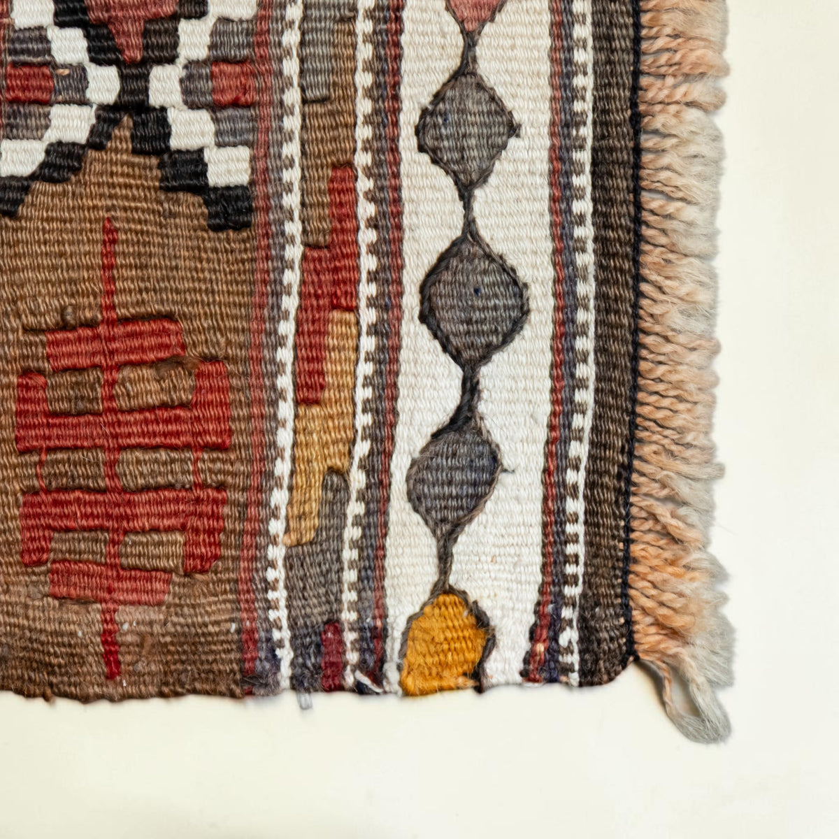 Close-up of a woven textile corner showing geometric patterns in brown, red, cream, gray, black, and mustard yellow with fringed edges on a light neutral background.