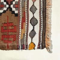 Close-up of a woven textile corner showing geometric patterns in brown, red, cream, gray, black, and mustard yellow with fringed edges on a light neutral background.