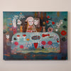 A colorful fantasy-style painting on canvas depicting a surreal table scene with stylized faces, animals, flowers, and abstract elements, hung on a plain light wall.