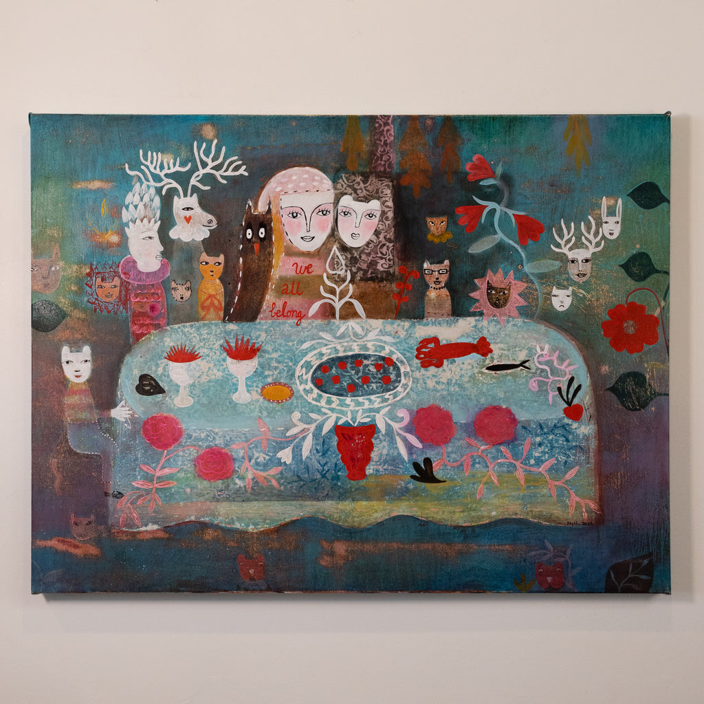 A colorful fantasy-style painting on canvas depicting a surreal table scene with stylized faces, animals, flowers, and abstract elements, hung on a plain light wall.