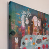 A square canvas painting shown at an angle against a plain light wall. The artwork features stylized faces, animals, and abstract forms in teal, blue, brown, white, pink, red, and orange tones.