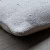 A close-up of a textured off-white sheepskin or wool throw pillow resting on a dark wood floor.