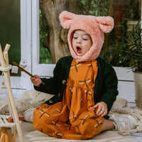 Child wearing a pink hooded coat with bear ears, sitting on a blanket indoors.