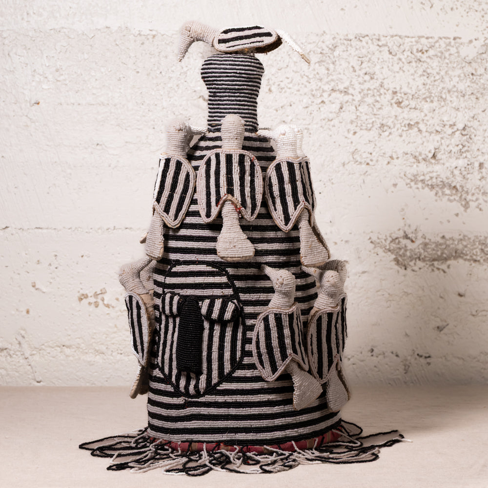 A handmade textile sculpture of a rooster, featuring black and white striped fabric with beige feather details, set against a textured off-white brick wall.
