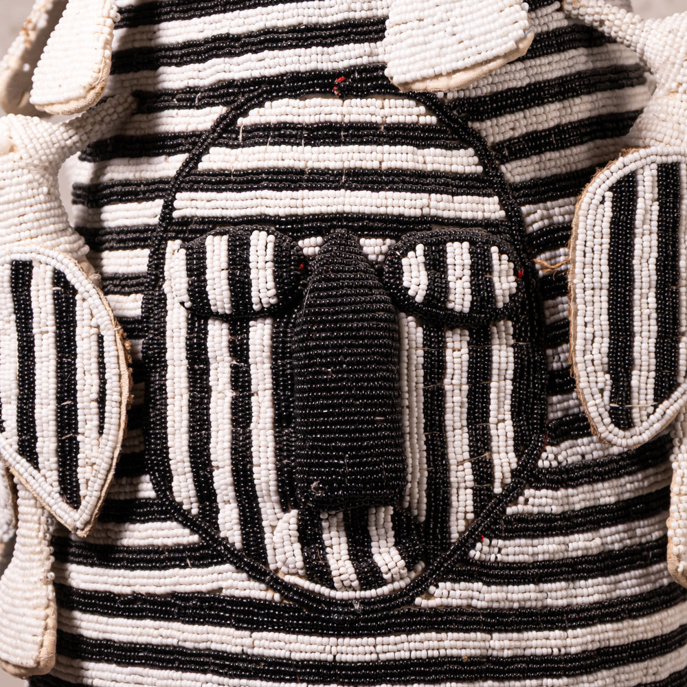 Close-up of a hand-beaded textile or bag featuring a stylized face design with black and off-white beadwork, striped horizontal bands, and leaf-like side motifs.