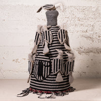 A handmade textile doll or decorative figure with black and white striped fabric, beige felted wool details, and hanging tassels, set against a textured off-white wall.