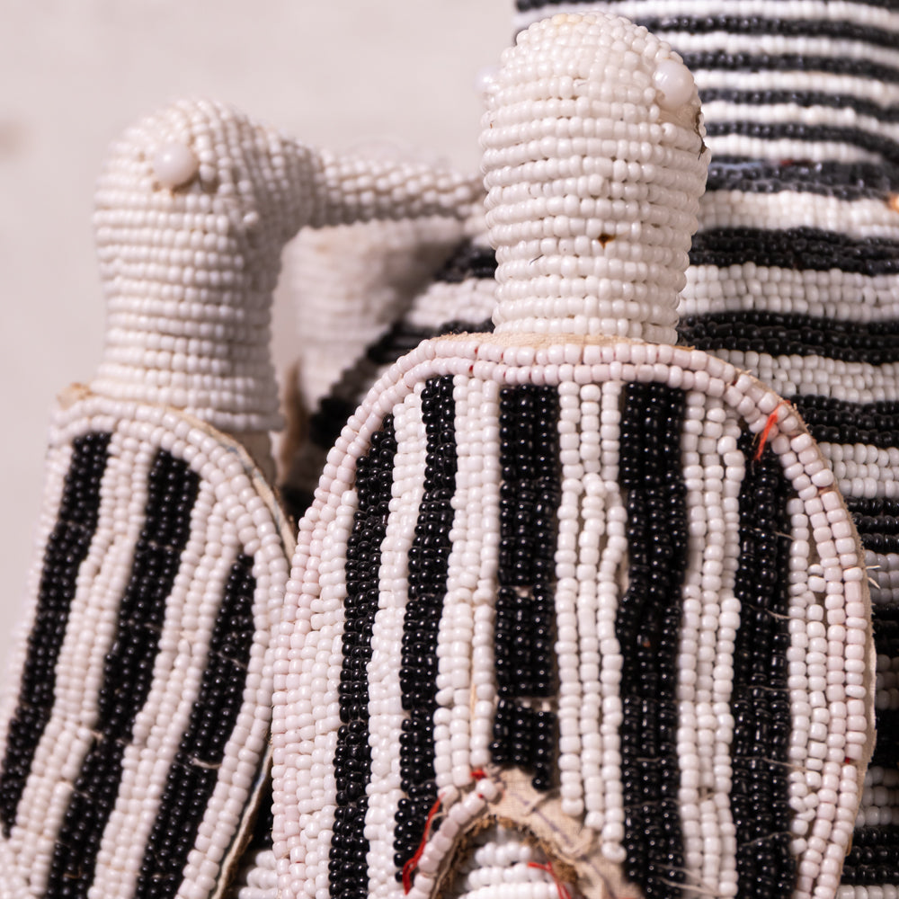 Close-up of hand-beaded African dolls, featuring black and white striped bodies made of small seed beads, with one doll in the foreground and another partially visible behind it.