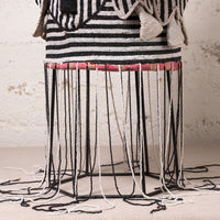 A close-up of a woven textile or rug with horizontal black and white stripes, draped over a black metal hexagonal frame with long hanging black and white cords. A pink patterned band is visible around the upper section, and the background is a textured off-white wall.