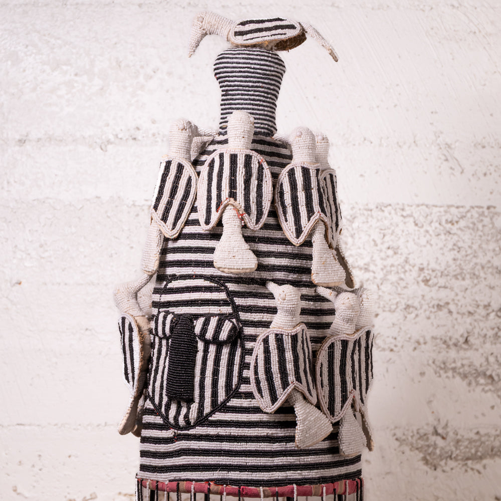 A handmade African-style textile doll or sculpture with black and white striped fabric and cream-colored fabric bird figures attached, set against a textured white wall.