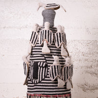 A handmade African-style textile doll or sculpture with black and white striped fabric and cream-colored fabric bird figures attached, set against a textured white wall.