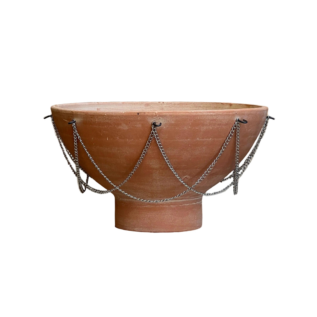 A terracotta clay pedestal bowl with draped silver metal chains, set against a solid beige background.