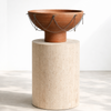 A terracotta clay bowl with hanging metal chains, displayed on a light beige stone pedestal against a clean white studio background.