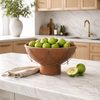 A large round terracotta bowl filled with green apples sits on a white marble kitchen island, with a folded beige napkin and two loose green apples beside it. The background shows a softly blurred modern kitchen with light wood cabinets, marble backsplash, and a brass faucet.