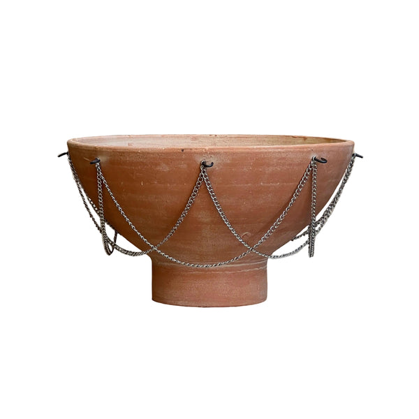 A terracotta clay pedestal bowl with draped silver metal chains, set against a solid beige background.