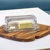 Clear glass butter dish with a wooden cutting board on a white surface.