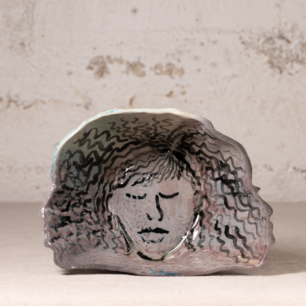 A handmade ceramic dish with a hand-painted face and wavy hair design, displayed on a neutral surface against a textured plaster wall.