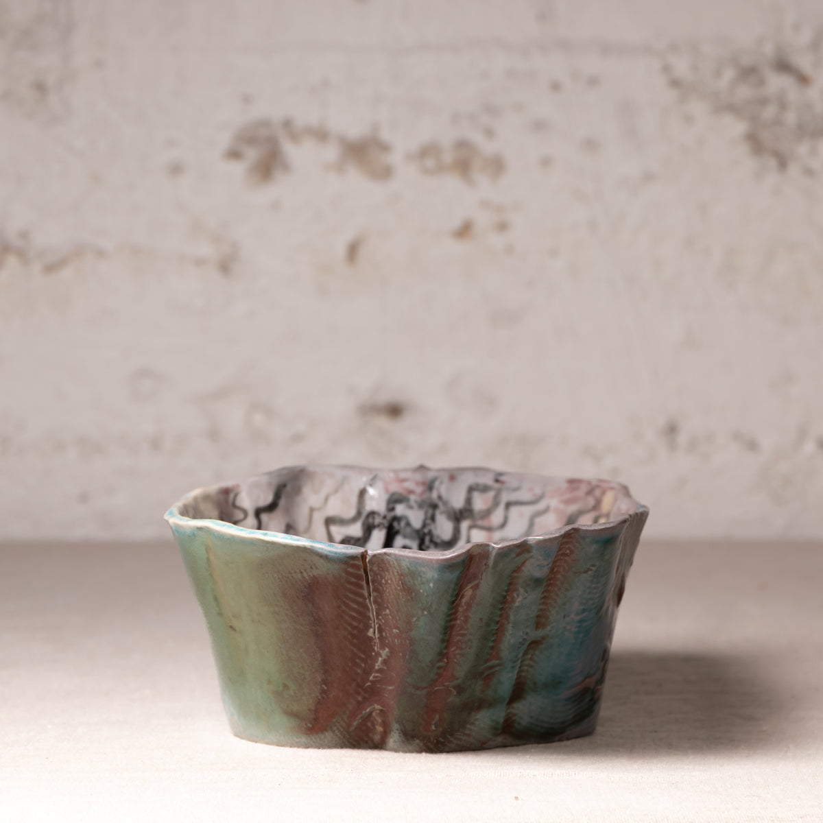 A handmade ceramic bowl with a wavy, scalloped rim and a marbled glaze of teal, green, brown, and black, set against a rustic, textured off-white brick wall background.