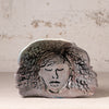 A handmade ceramic dish with a hand-painted face and wavy hair design, displayed on a neutral surface against a textured plaster wall.
