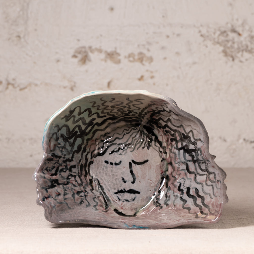 A handmade ceramic dish with a hand-painted face and wavy hair design, displayed on a neutral surface against a textured plaster wall.