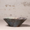 A handmade ceramic bowl with a textured, organic scalloped rim, featuring a dark blue-gray exterior with brown rust-like accents and a light green glazed interior, set against a rustic, light beige plaster wall.