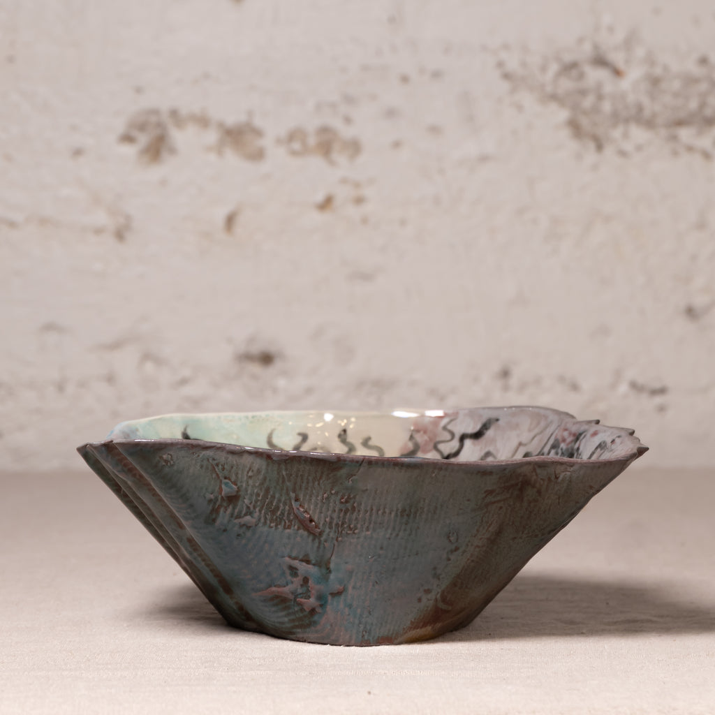 A handmade ceramic bowl with a textured, organic scalloped rim, featuring a dark blue-gray exterior with brown rust-like accents and a light green glazed interior, set against a rustic, light beige plaster wall.