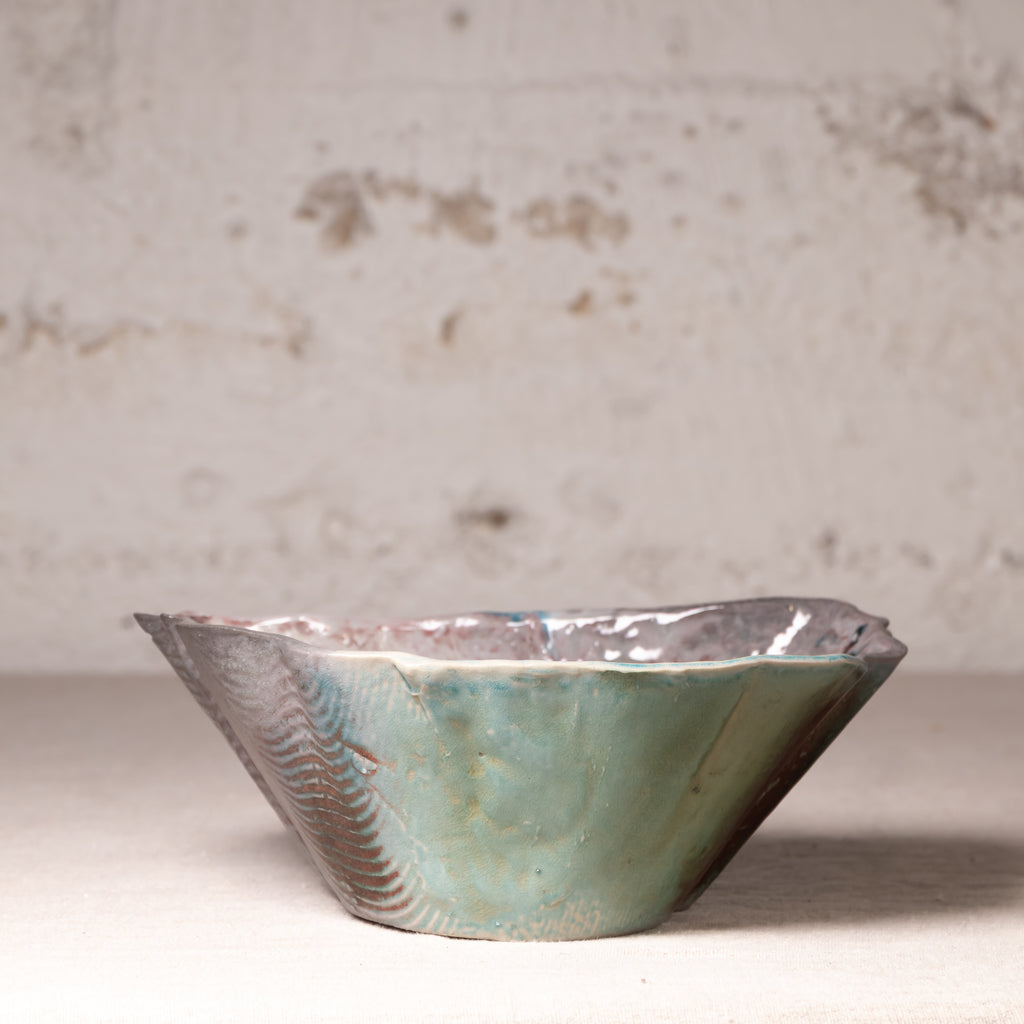 A handmade ceramic bowl with a wavy, irregular rim, featuring a textured sage green glaze on the front and a metallic silver patterned exterior, set against a rustic, distressed off-white wall.