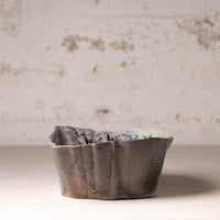 A handmade ceramic bowl with a textured, organic scalloped rim and a marbled earthy glaze, set against a rustic, distressed plaster wall.