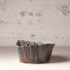 A handmade ceramic bowl with a textured, organic scalloped rim and a marbled earthy glaze, set against a rustic, distressed plaster wall.