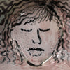 A close-up of a textured ceramic bowl featuring a stylized, hand-painted face with closed eyes in black and muted pink tones.