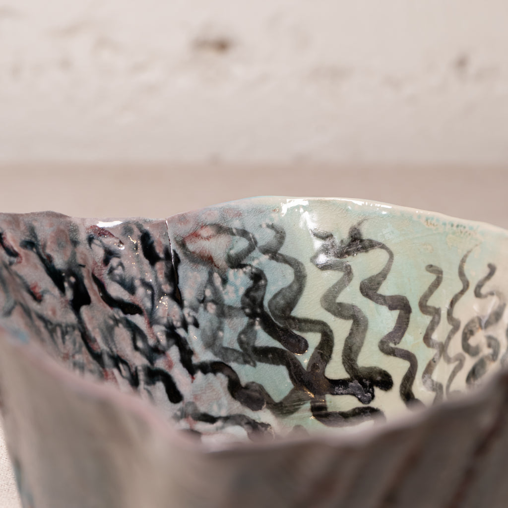 Close-up of a handmade ceramic bowl with a textured, organic wavy pattern in black, gray, and muted blue-green glaze, set against a neutral beige background.