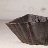 A close-up of a handmade ceramic bowl with a dark, textured glaze and organic, wavy edges, set against a neutral, light beige background.