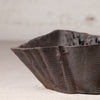 A close-up of a handmade ceramic bowl with a dark, textured glaze and organic, wavy edges, set against a neutral, light beige background.