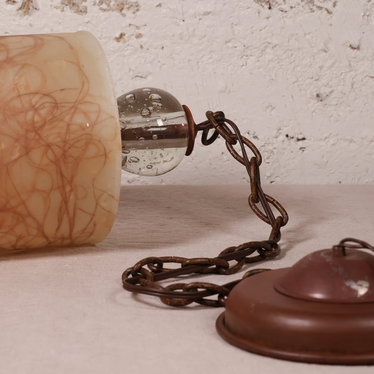 A close-up of a vintage-style hanging lamp or lantern component with a translucent beige marbled glass shade, a clear glass bulb, a dark metal chain, and a reddish-brown metal cap against a textured off-white wall.