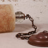 A close-up of a vintage-style hanging lamp or lantern component with a translucent beige marbled glass shade, a clear glass bulb, a dark metal chain, and a reddish-brown metal cap against a textured off-white wall.