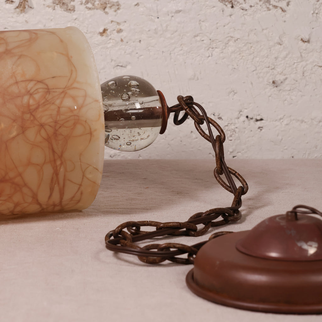 A close-up of a vintage-style hanging lamp or lantern component with a translucent beige marbled glass shade, a clear glass bulb, a dark metal chain, and a reddish-brown metal cap against a textured off-white wall.