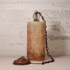 A vintage-style table lamp with a cylindrical beige and brown marbled stone or resin body, a dark metal chain, a clear glass globe bulb, and a round dark metal base, set against a textured off-white brick wall.