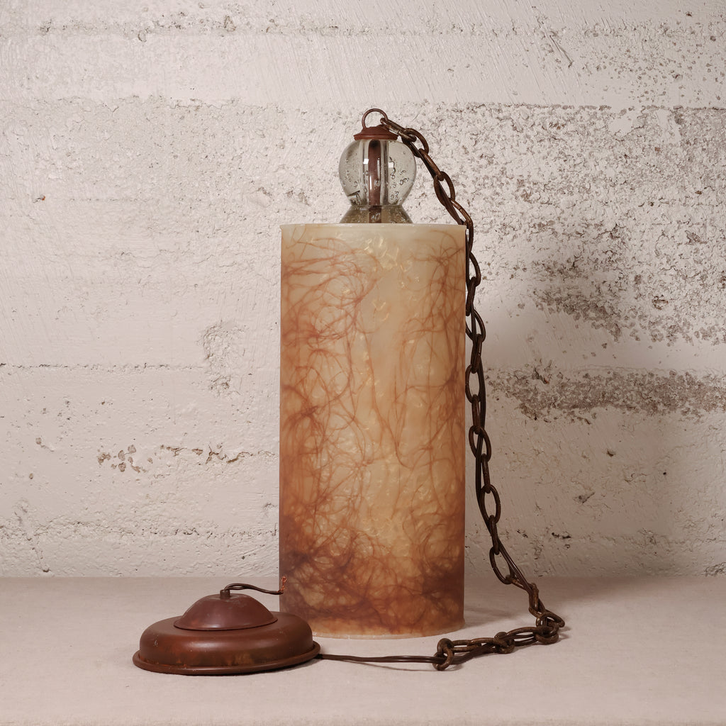 A vintage-style table lamp with a cylindrical beige and brown marbled stone or resin body, a dark metal chain, a clear glass globe bulb, and a round dark metal base, set against a textured off-white brick wall.