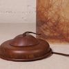 A close-up of a vintage-style metal lamp base or switch component in a dark reddish-brown finish, with a thin wire loop and visible exposed copper terminals, set against a textured off-white background with part of a brown marbled object visible behind it.