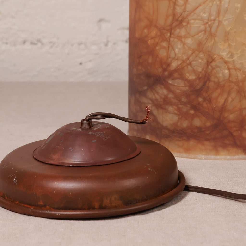 A close-up of a vintage-style metal lamp base or switch component in a dark reddish-brown finish, with a thin wire loop and visible exposed copper terminals, set against a textured off-white background with part of a brown marbled object visible behind it.