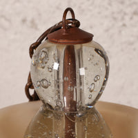 Close-up of a hanging glass lantern with a clear bubble glass body, copper-toned metal top and chain, and a beige shade below.