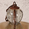 Close-up of a hanging glass lantern with a clear bubble glass body, copper-toned metal top and chain, and a beige shade below.