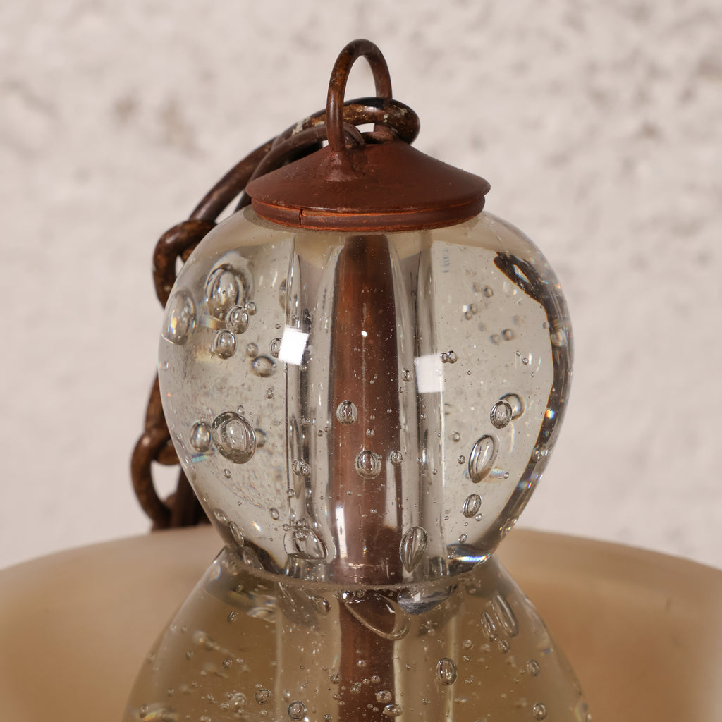 Close-up of a hanging glass lantern with a clear bubble glass body, copper-toned metal top and chain, and a beige shade below.