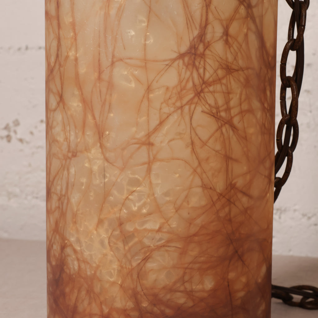 A close-up of a cylindrical pendant light with a textured, translucent amber-brown glass shade filled with a dense network of dark brown hair, with a dark metal chain visible on the right side.