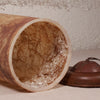 A close-up of a cylindrical lampshade or decorative container lying on its side, showing a brown marbled exterior and a clear, textured interior filled with dark fibers. A matching brown metal cap or lid is placed beside it on a light surface against a textured beige wall.