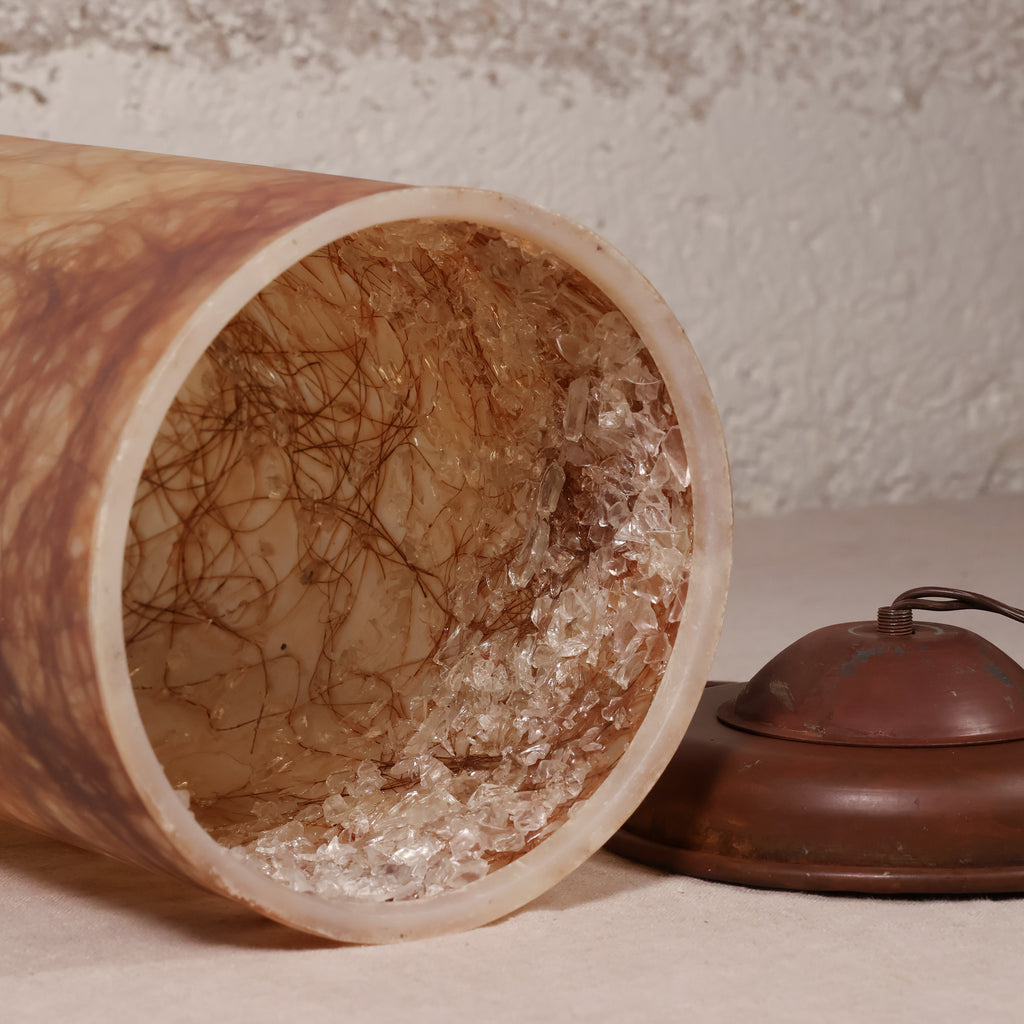 A close-up of a cylindrical lampshade or decorative container lying on its side, showing a brown marbled exterior and a clear, textured interior filled with dark fibers. A matching brown metal cap or lid is placed beside it on a light surface against a textured beige wall.